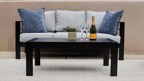 A black coffee table with a bottle of champagne, two glasses, and a small plant on a light grey sofa with blue cushions.