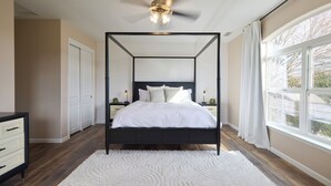 A four-poster bed with a ceiling fan, white bedding, and a view of the outdoors through a large window.