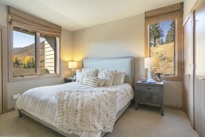 Una camera da letto con un letto grande, due comodini con lampade, un tavolino e vista su un paesaggio montuoso dalle finestre.