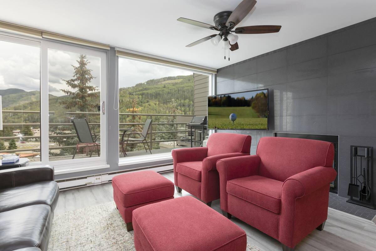 A modern living room with a large TV, red armchairs, a sofa, and a balcony with a view of the mountains.