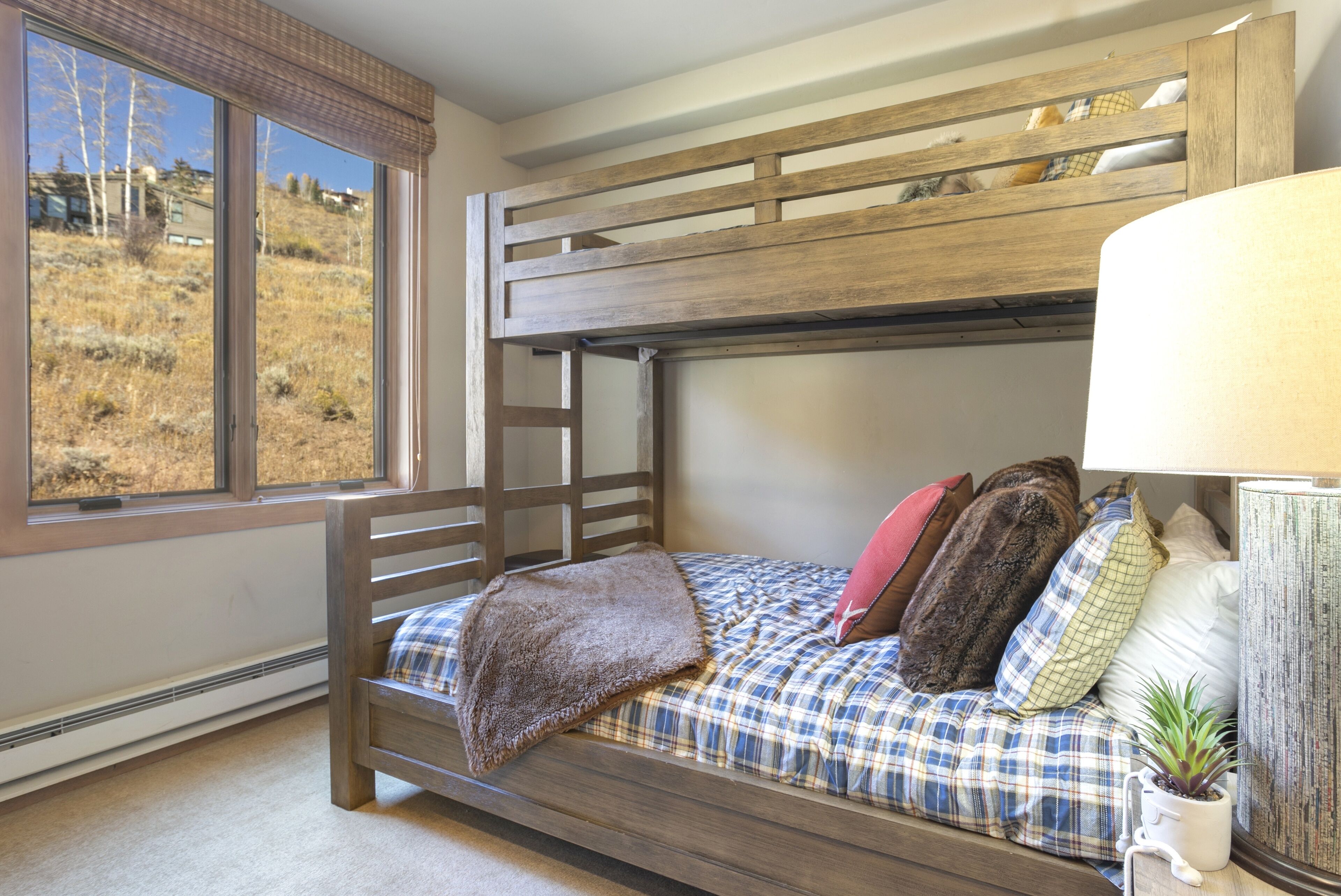 A bunk bed with a plaid bedspread and pillows, a window with a view of a hillside, and a bedside lamp.