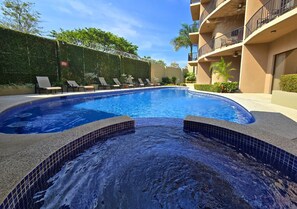 Une piscine avec un espace équipé d’un jacuzzi, entourée de chaises longues et bordée d’un bâtiment doté de balcons.