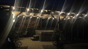 An outdoor patio area with a pergola covered in string lights, a hot tub, and a seating area with chairs.