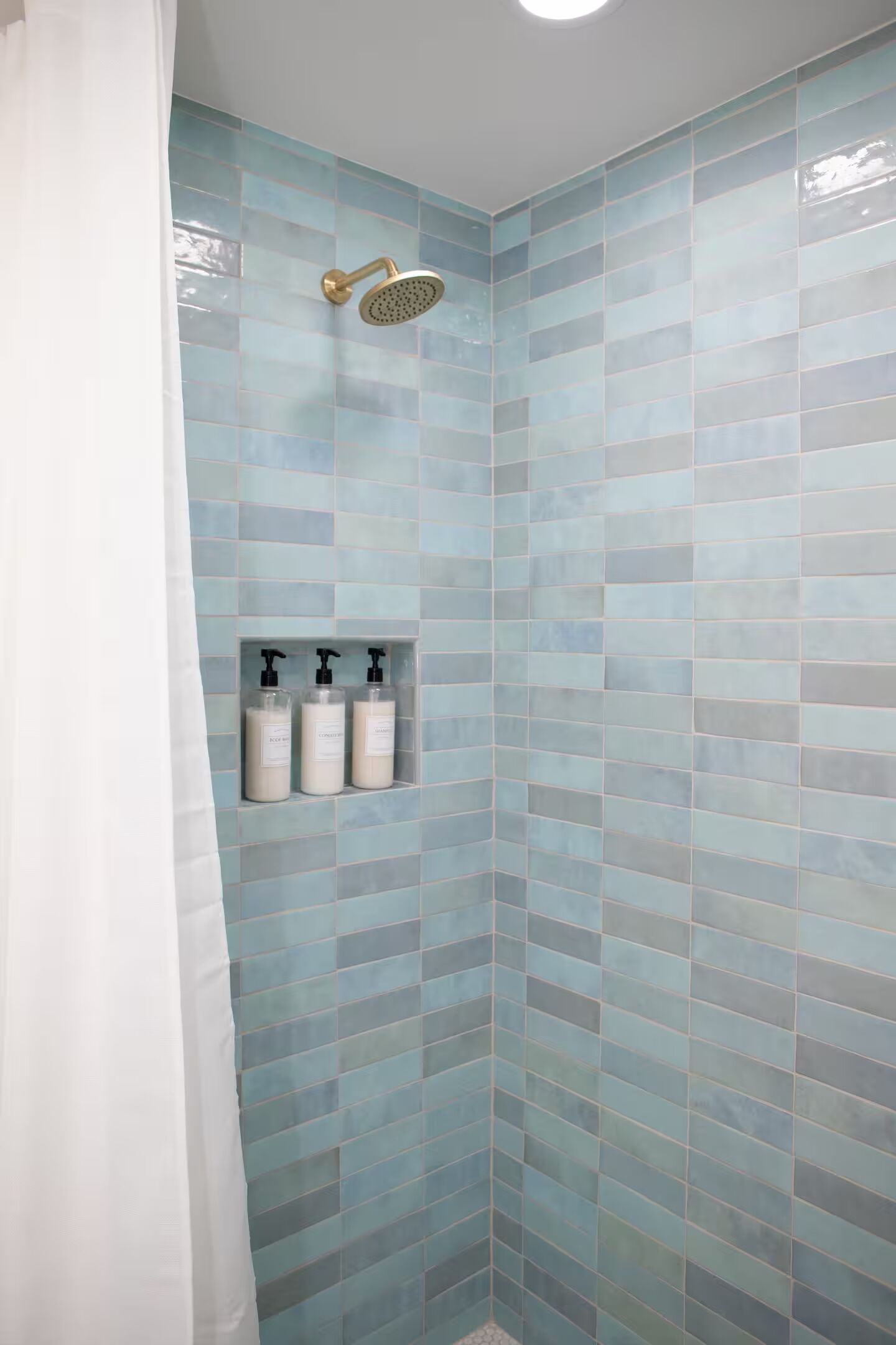 Une douche aux murs recouverts de carreaux bleus, un pommeau de douche doré et trois bouteilles de shampoing sur une étagère.