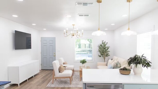 A modern kitchen with white cabinetry, a built-in oven, and a stainless steel dishwasher. There is a dining area with a white table and chairs, and a large window providing natural light.