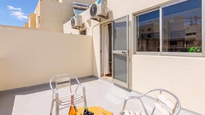 Un balcon avec deux chaises en métal, une table avec une bouteille et des fruits, et des unités de climatisation fixées au mur.