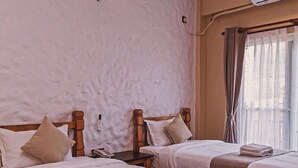 A hotel room with two beds, a wall-mounted air conditioner, a bedside table with a lamp, and a window with curtains. - Cozy Home (Pokhara)