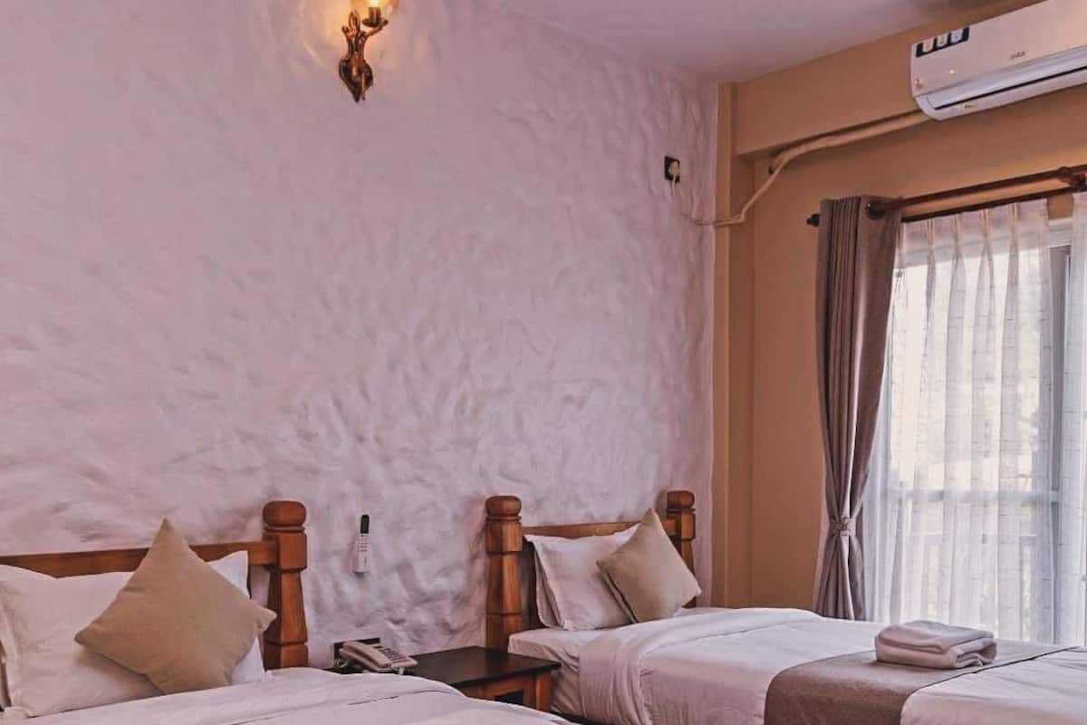 Une chambre d’hôtel avec deux lits, un climatiseur fixé au mur, une table de chevet avec une lampe et une fenêtre avec des rideaux.
