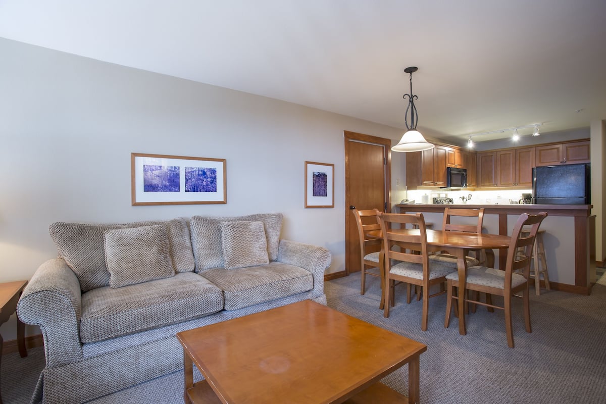 A living room with a sofa, a coffee table, and a dining area with chairs and a table. There is a kitchen area with a refrigerator, microwave, and a TV.