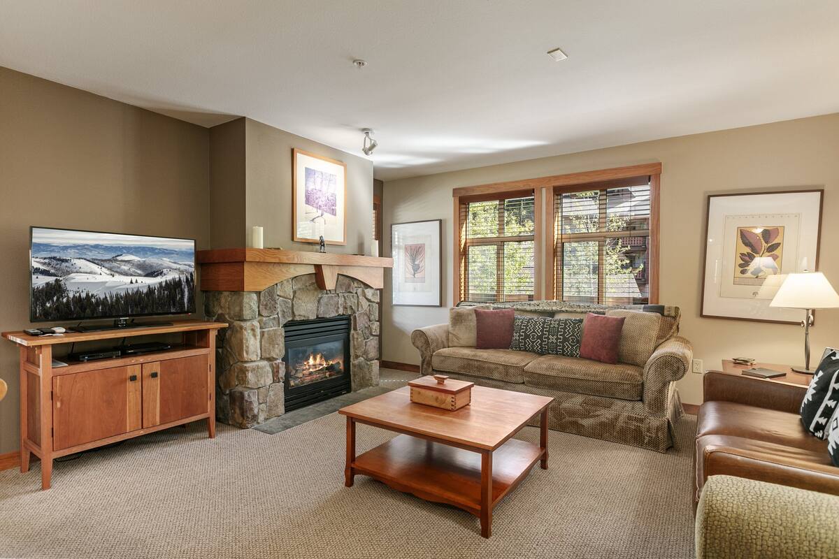 A cozy living room with a stone fireplace, a large flat-screen TV, a wooden coffee table, and a comfortable sofa.