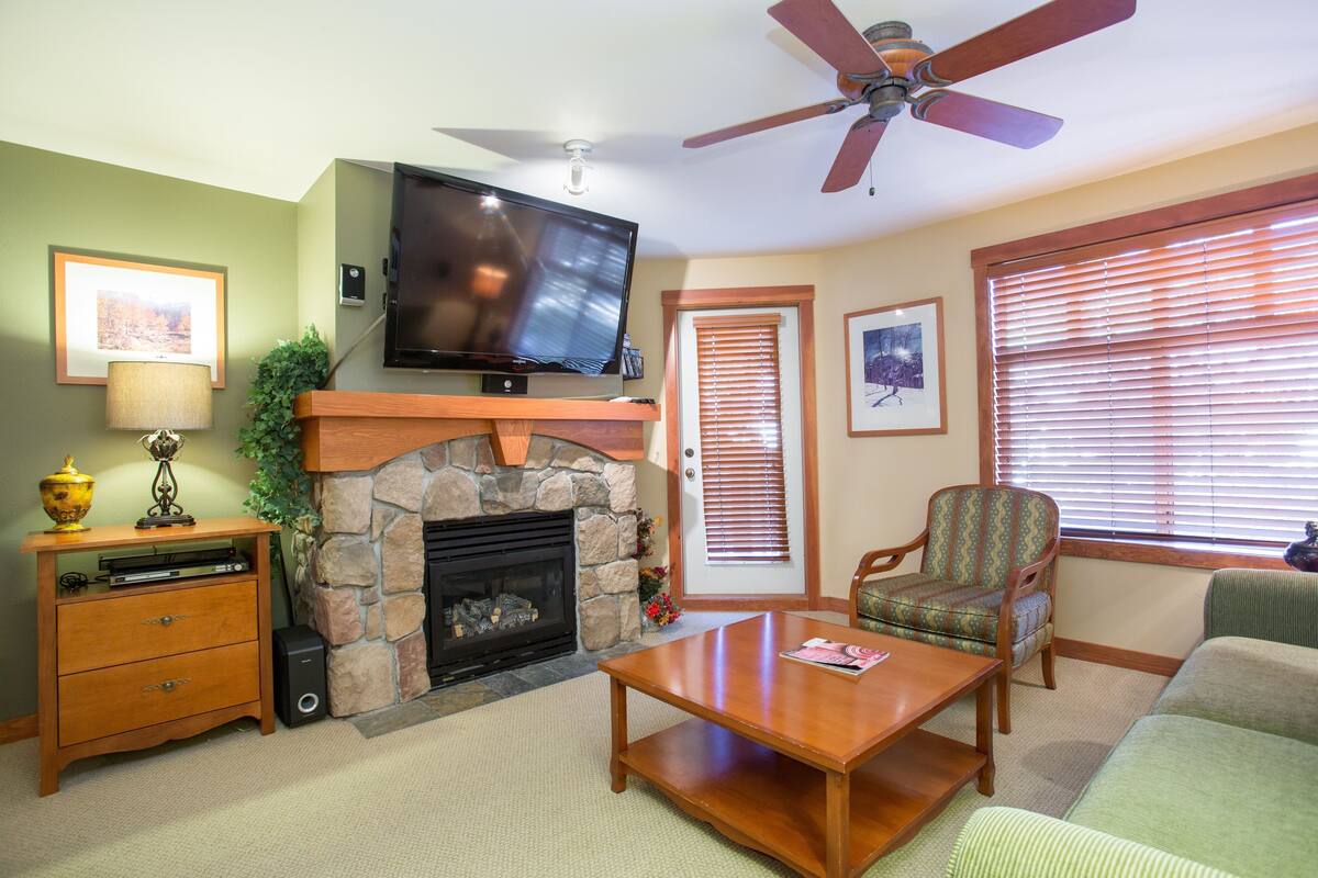 A living room with a stone fireplace, a television mounted above it, a ceiling fan, and a window with blinds.
