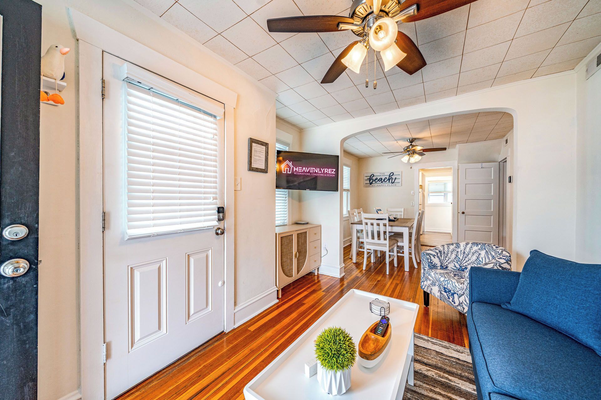 A spacious living room with a wooden floor, a ceiling fan, a TV mounted on the wall, and a dining area with a table and chairs.