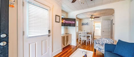A spacious living room with a wooden floor, a ceiling fan, a TV mounted on the wall, and a dining area with a table and chairs.
