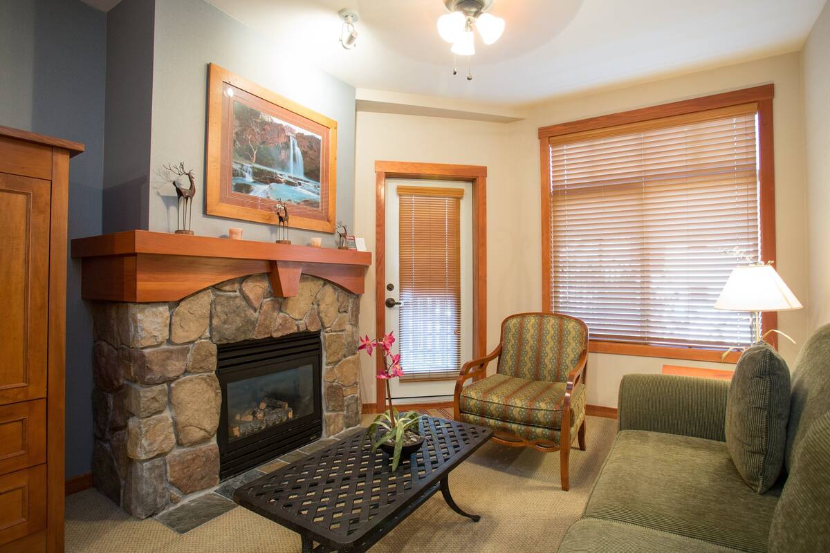 A living room with a stone fireplace, a framed picture above it, a green armchair, a sofa, and a window with blinds.