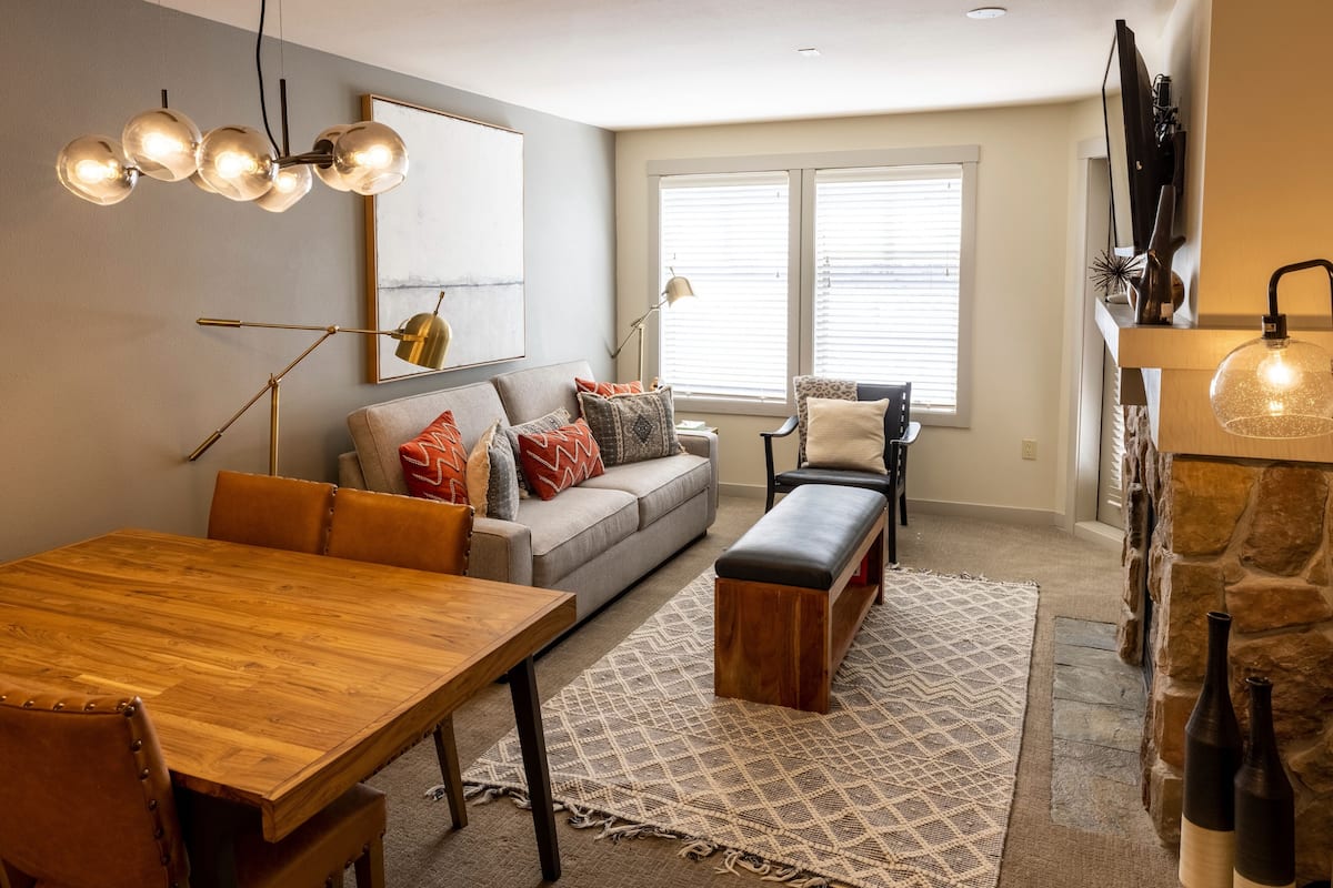 A modern living room with a wooden dining table, a gray sofa with patterned cushions, a bench, and a stone fireplace.