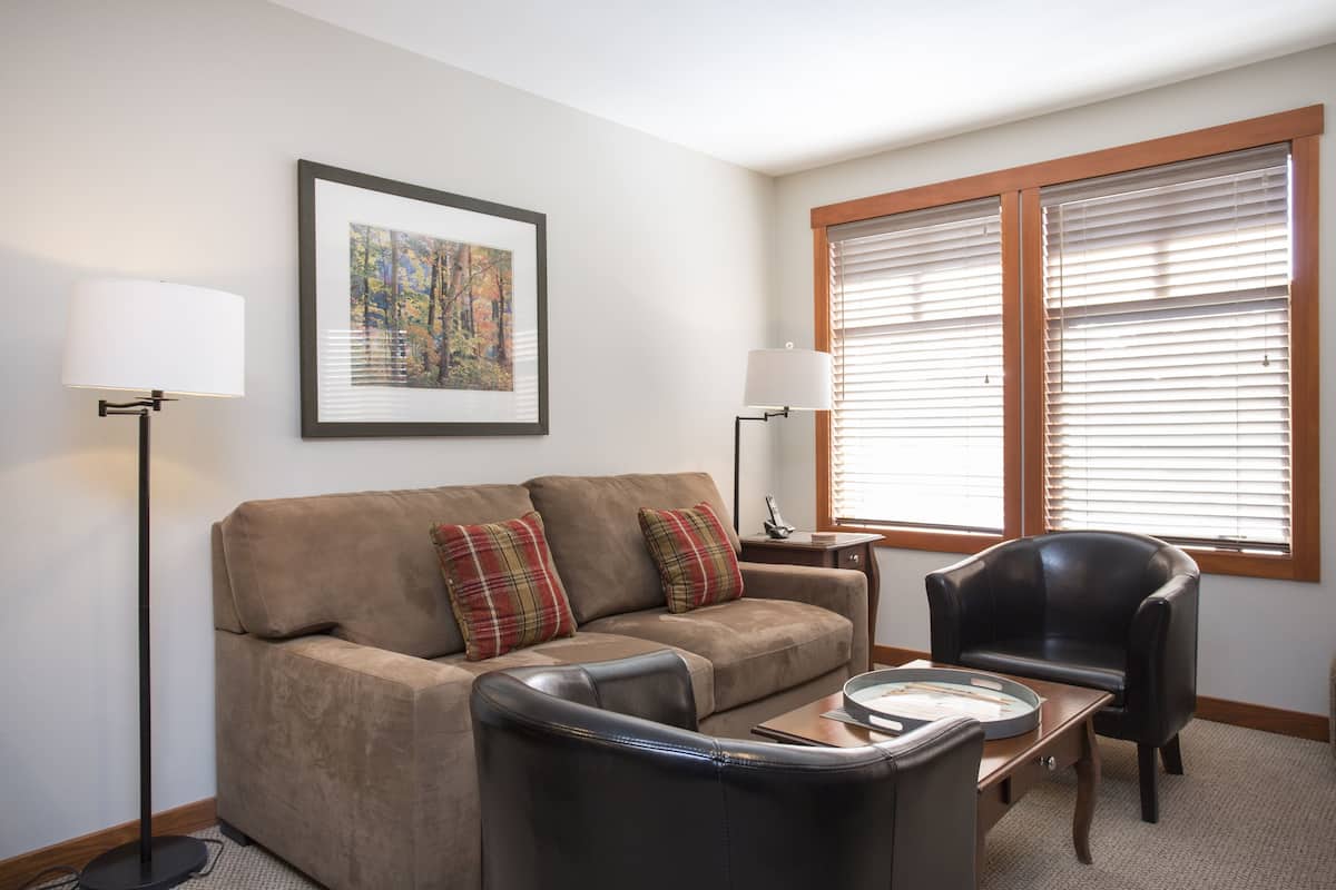 A living room with a beige sofa, two plaid cushions, a black leather chair, a coffee table, a floor lamp, and a framed picture on the wall.