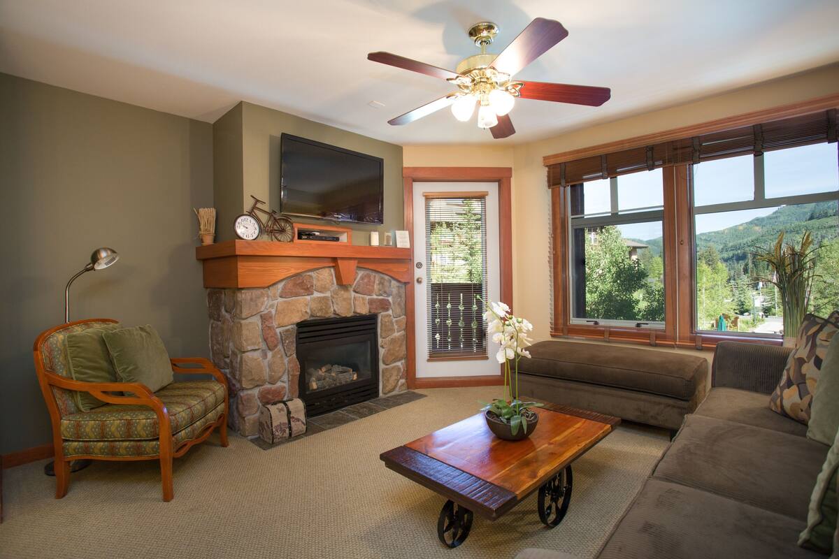 A living room with a stone fireplace, a television, a sofa, a coffee table, and a view of the outdoors.