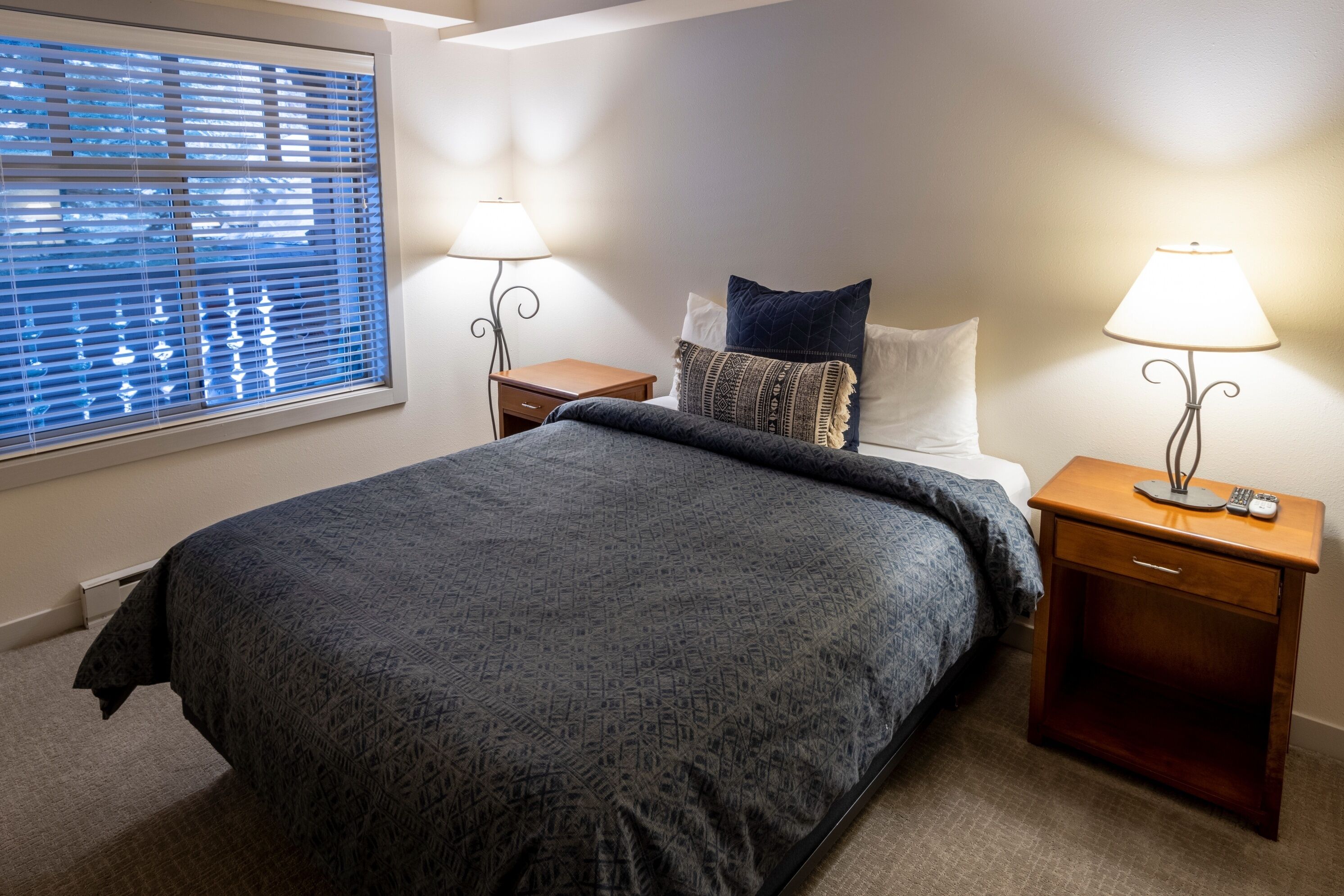 A neatly arranged bedroom with a large bed, two bedside tables with lamps, a window with blinds, and a bedside drawer.