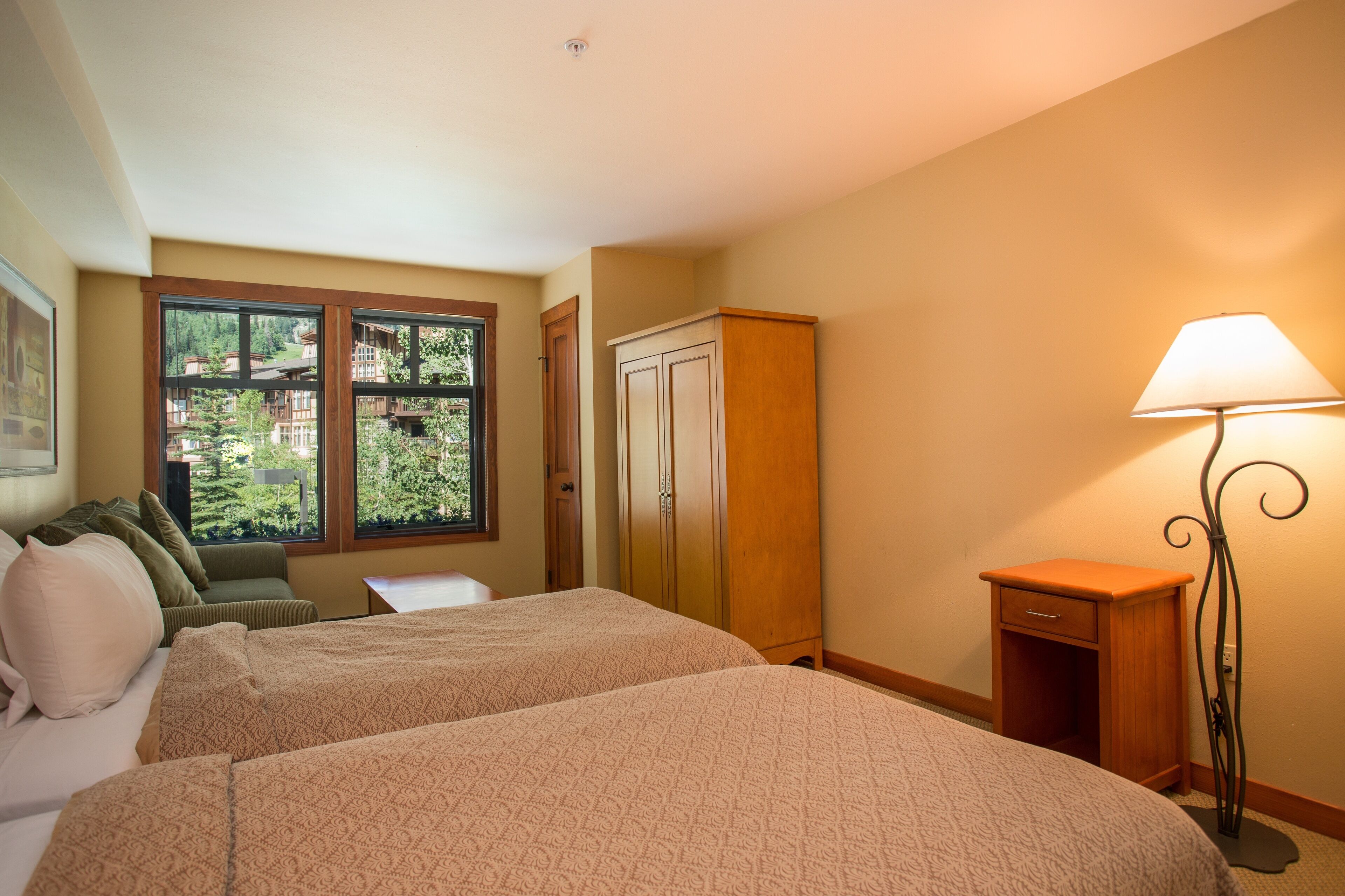 Quarto de hotel com duas camas, janela com vista para o verde, guarda-roupa de madeira e mesa de cabeceira com abajur.