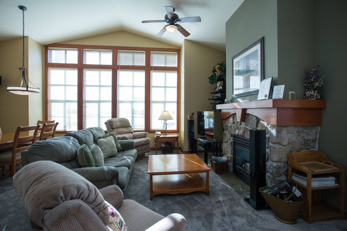 A living room with a fireplace, a television, and a large window with blinds.