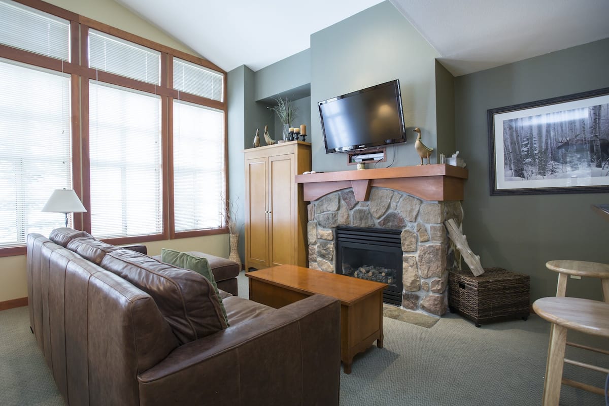 A living room with a stone fireplace, leather sofa, wooden coffee table, and a television mounted above the fireplace.