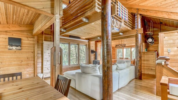 A wooden cabin interior with a dining area, a sofa, and a large window.