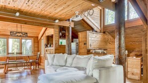 A cozy cabin interior with a large sectional sofa, wooden walls, and a dining area with a table and chairs.