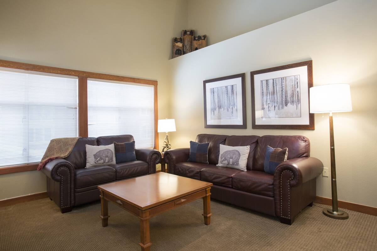 A living room with two brown leather sofas, a wooden coffee table, and a floor lamp. There are two framed pictures on the wall and a window with blinds.