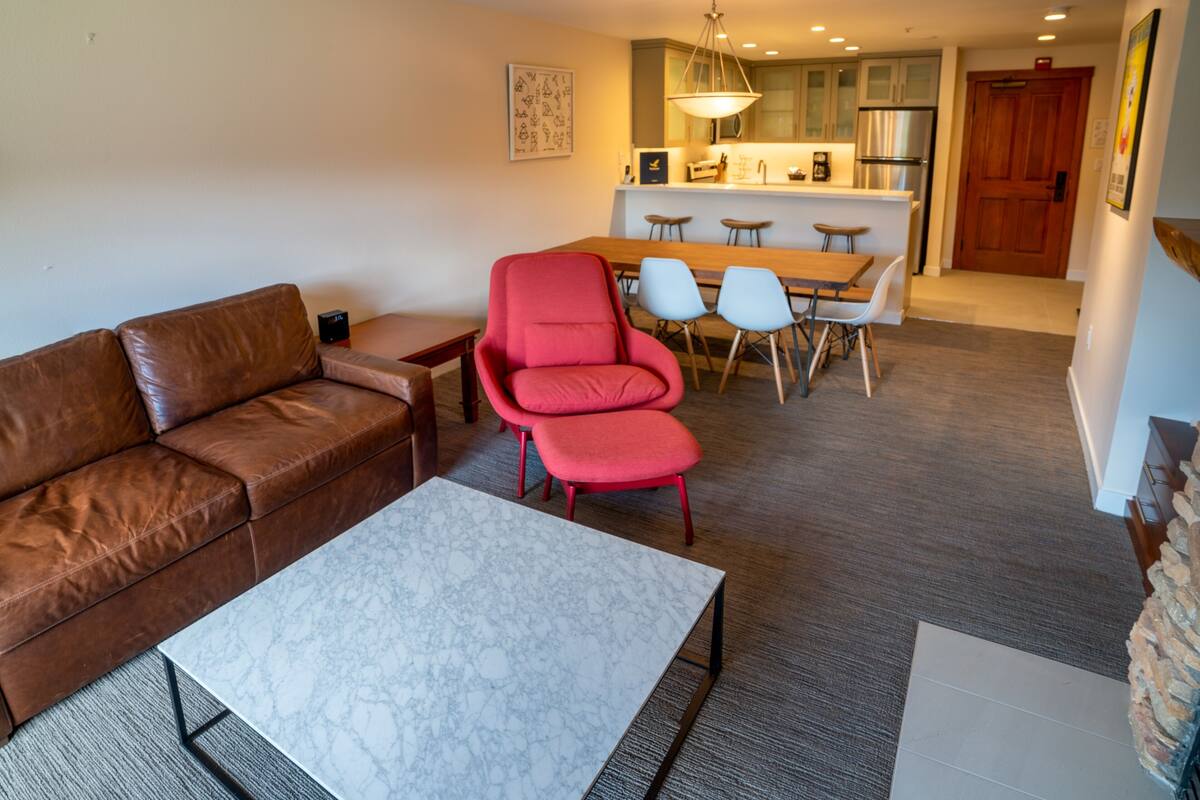 A living room with a brown leather sofa, a marble coffee table, and a red chair. There is a dining area with a wooden table and chairs. The kitchen is visible in the background with modern appliances.