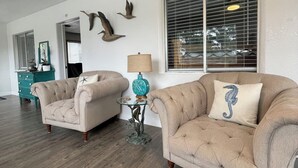 A spacious living room with two beige armchairs, a glass coffee table, and a teal side table with a lamp.