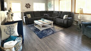 A living room with a wooden ceiling, a ceiling fan, a large window, a grey sofa, a coffee table, and a blue rug.