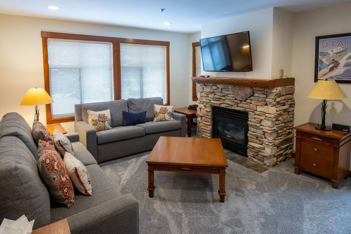 A living room with a stone fireplace, a flat-screen TV mounted above it, a grey sofa with patterned cushions, a wooden coffee table, and a lamp on a wooden side table.