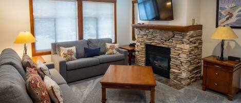 A living room with a stone fireplace, a flat-screen TV mounted above it, a grey sofa with patterned cushions, a wooden coffee table, and a lamp on a wooden side table.