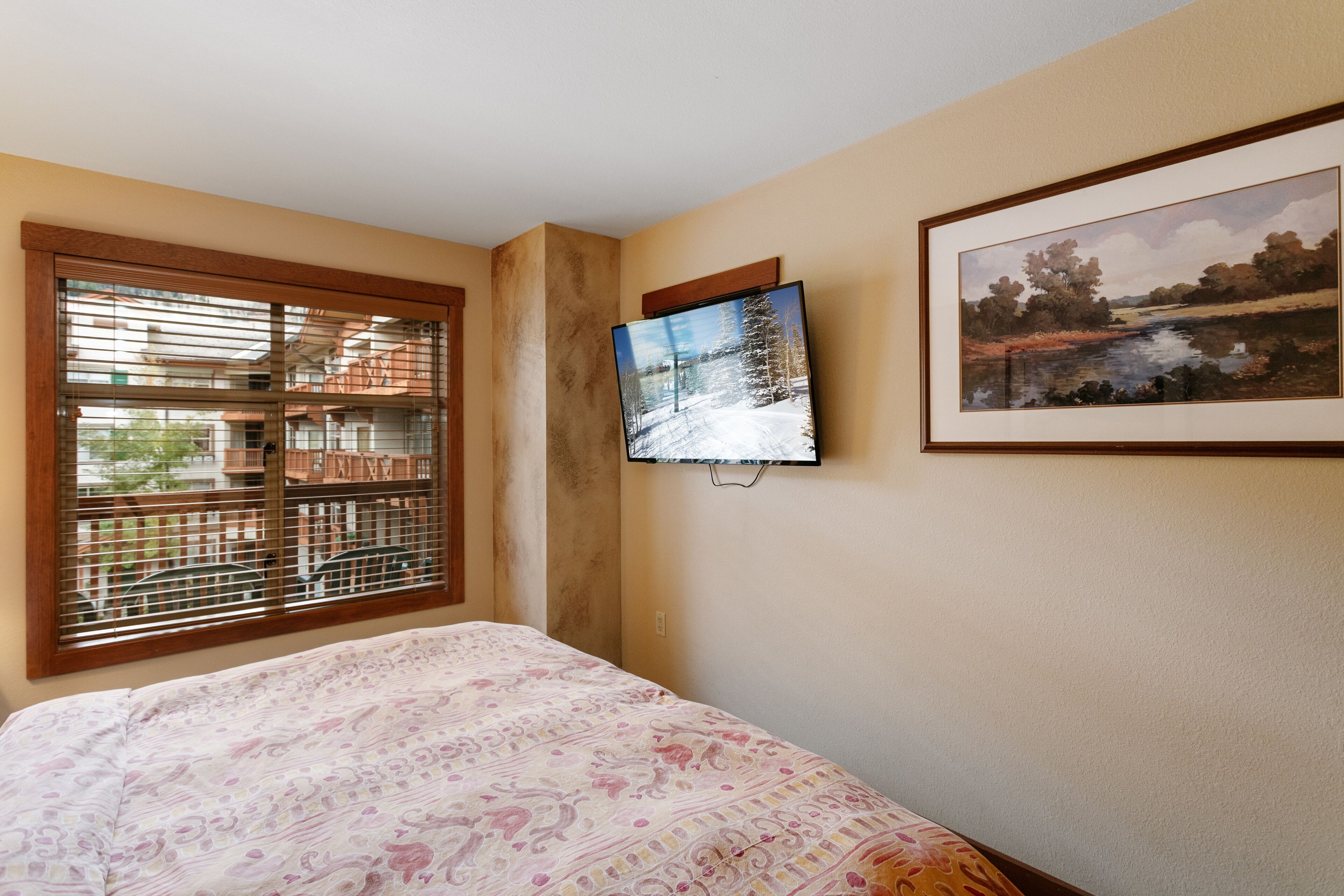 Un dormitorio con cama, ventana con persianas, televisor de pantalla plana montado en la pared y un cuadro enmarcado con un paisaje.