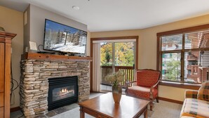 A living room with a stone fireplace, a television mounted above it, a comfortable red chair, and a sofa. - Powderhorn Lodge Cozy Condos - Solitude UT (Solitude)