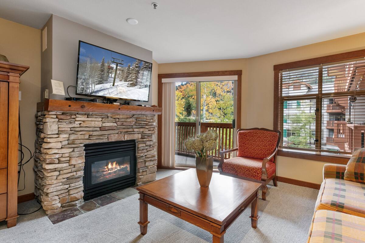 A living room with a stone fireplace, a flat-screen TV mounted above it, a wooden coffee table, a red patterned armchair, and a view of a balcony with autumn foliage.