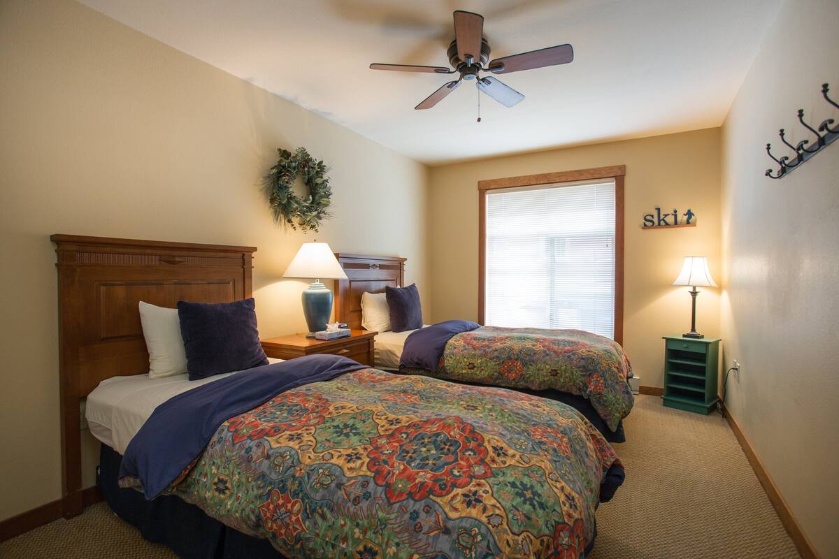 A bedroom with two beds, a ceiling fan, a window with blinds, a nightstand with a lamp, and a wall-mounted sign that says "ski".