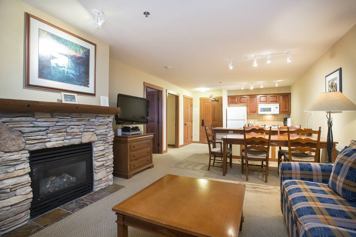 A living room with a stone fireplace, a television, a dining area with wooden chairs and a table, and a kitchen in the background.