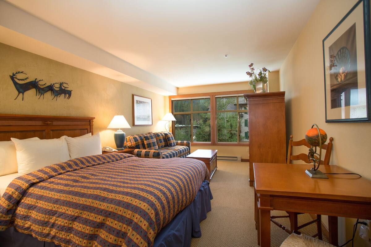 A hotel room with a large bed, a desk, and a chair. There is a window with a view of trees and a wall-mounted deer decoration.