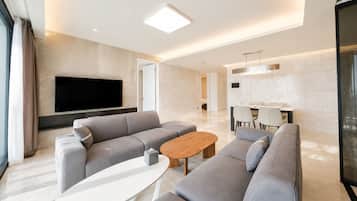 A modern living room with a large flat-screen TV, grey sofas, a wooden coffee table, and a dining area with chairs and a table.