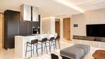 A modern kitchen with a central island, bar stools, a flat-screen TV, and a seating area with a sofa and ottoman.