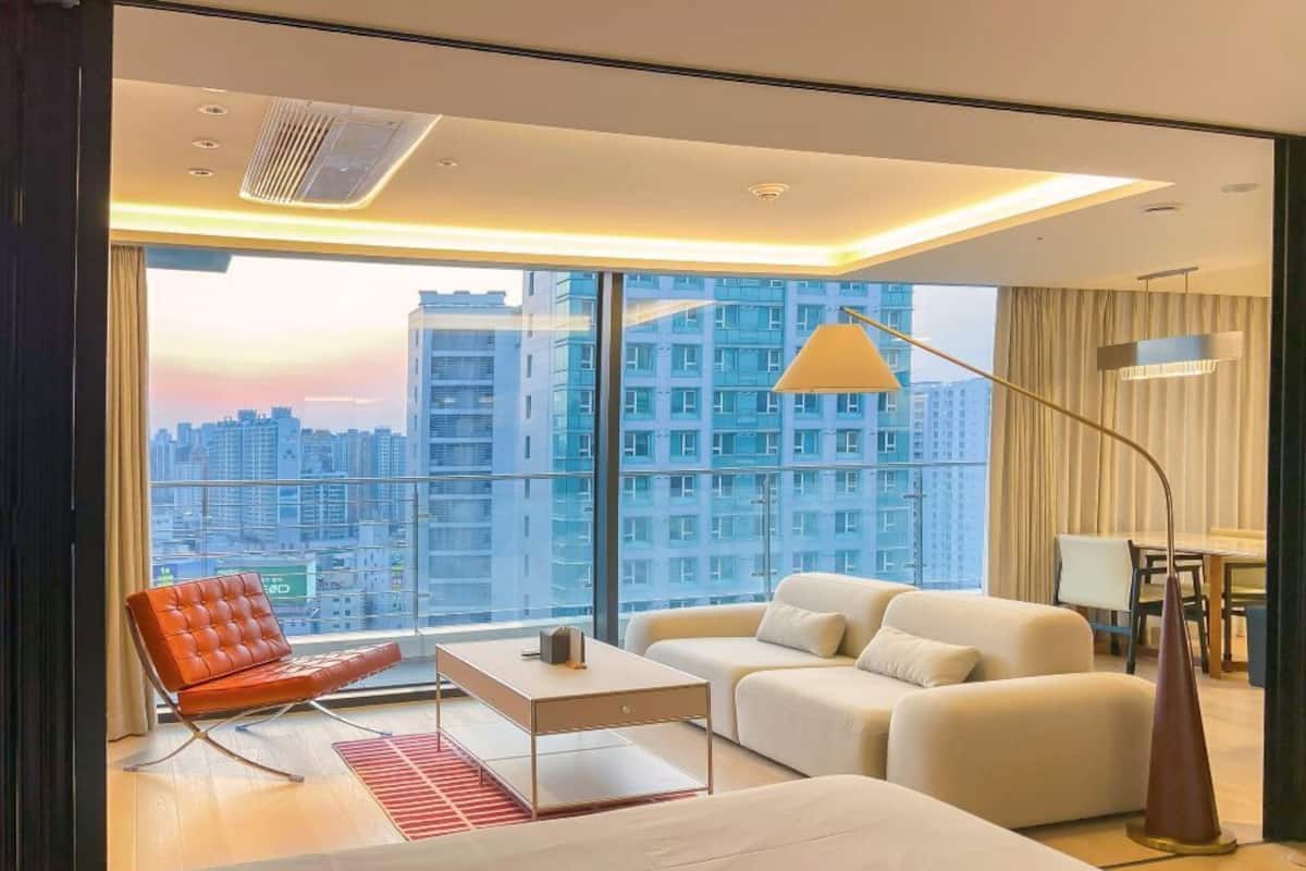 Une chambre d’hôtel moderne avec une grande fenêtre, un canapé, un fauteuil, une table et une lampe.