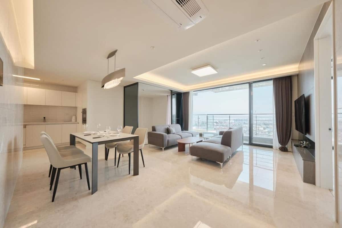 Une chambre d’hôtel moderne comprenant un coin repas, un espace de vie avec un canapé et des fauteuils, un téléviseur à écran plat et une grande fenêtre donnant sur la ville.