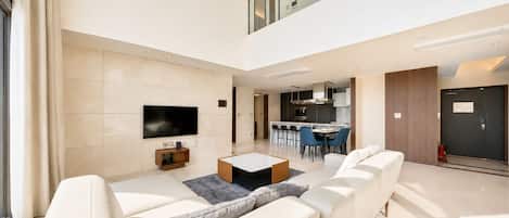 A modern living room with a flat-screen TV, a coffee table, and a sofa. There is a dining area with chairs and a kitchen in the background.