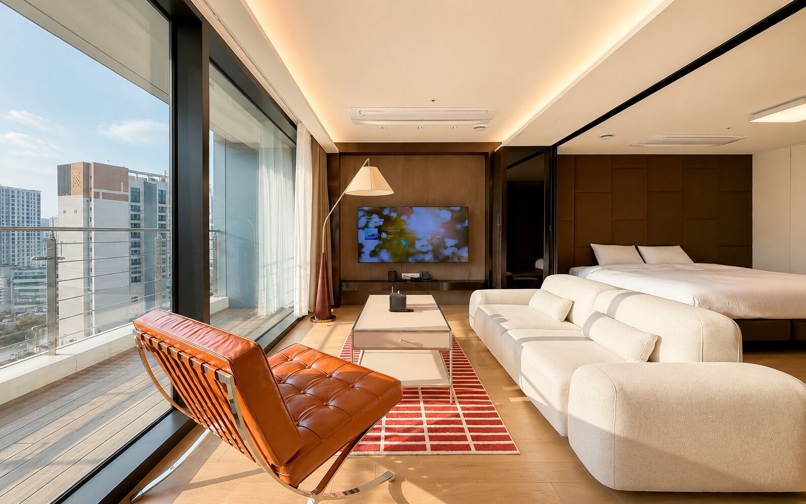 A modern hotel room with a large TV, a sofa, a bed, and a balcony with a view of the city.