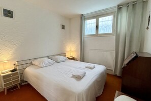 Habitación individual con una cama de soltera, cabecera blanca, lámparas de noche, ventana con cortinas, radiador y un escritorio con lámpara.