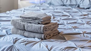 A neatly made bed with a grey duvet and pillows, a stack of folded towels, and a bedside table with a lamp. - Peaceful and Quiet 4 Br house with Garage (Sacramento)