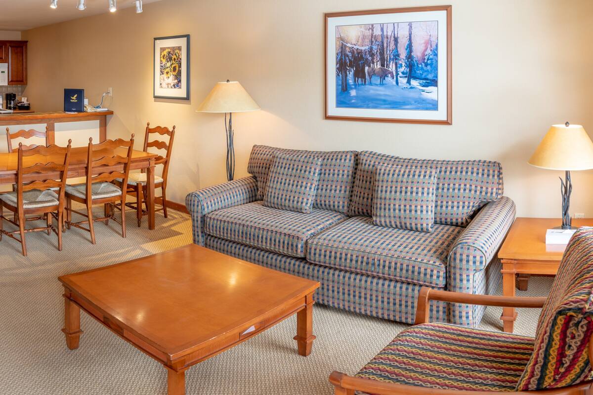 A living room with a patterned sofa, wooden coffee table, and matching chairs. There is a framed painting on the wall and a lamp on a side table.