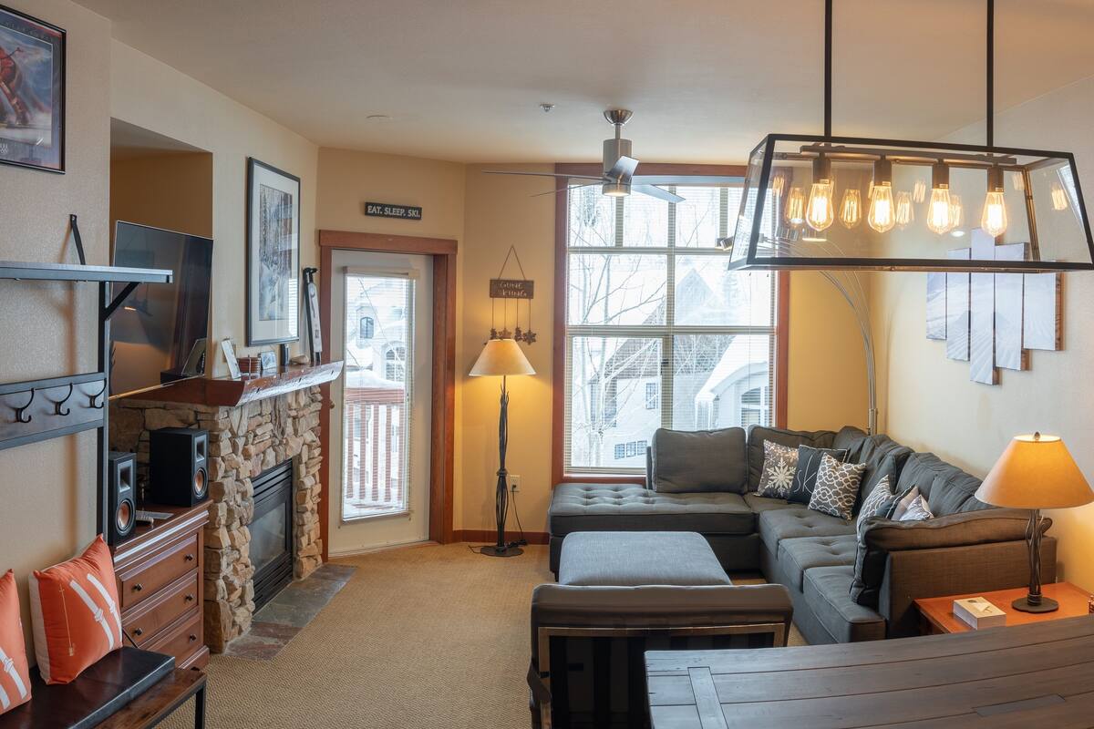 A cozy living room with a stone fireplace, a large window with blinds, a modern ceiling fan, and a comfortable sofa set.