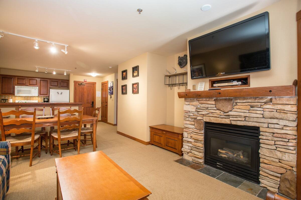 A spacious living room with a stone fireplace, a flat-screen TV mounted above it, and a dining area with wooden furniture.